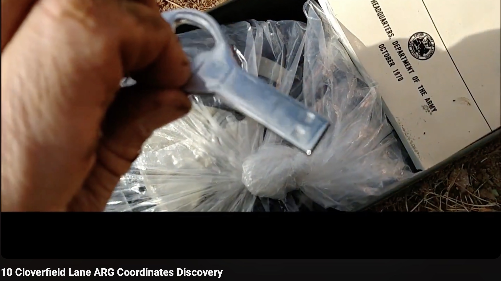 A hand reaching into a plastic bag to retrieve a small sealed container buried in dirt and straw, labeled with military-style coordinates text reading '10 Cloverfield Lane ARG Coordinates Discovery' — a physical cache planted as part of the ARG.