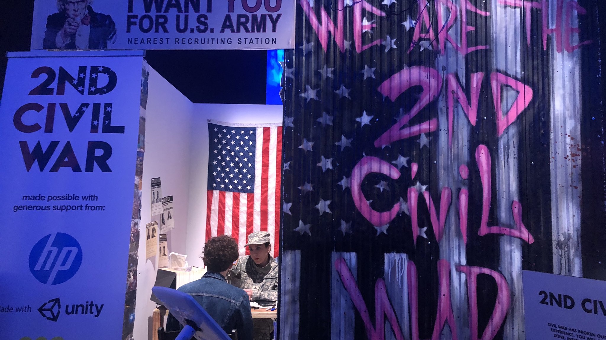 The 2nd Civil War VR installation at the 2019 Tribeca Film Festival, showing a street-art-covered installation booth with 'I Want You For U.S. Army' poster, an American flag, and graffiti reading 'We Take The 2nd Civil War,' with a visitor seated inside being briefed by a costumed actor.