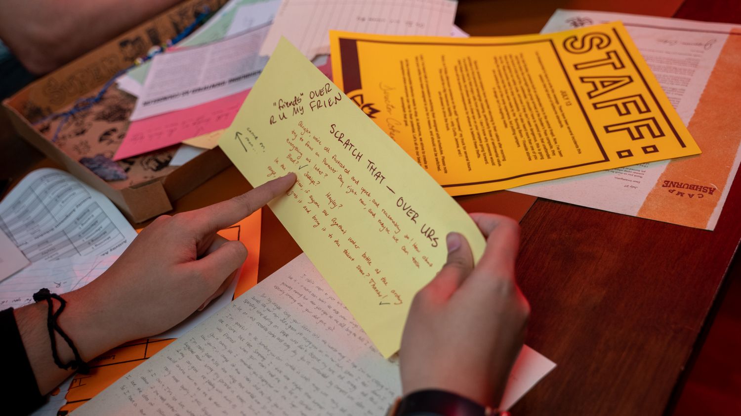 A hand holding a handwritten yellow sticky note over a spread of game materials including printed documents labeled 'Staff,' photographs, and colorful paper clues, suggesting active investigation gameplay.